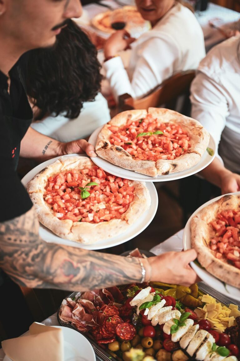 A waiter serves freshly baked Italian pizzas with an assortment of appetizers in a busy restaurant setting.