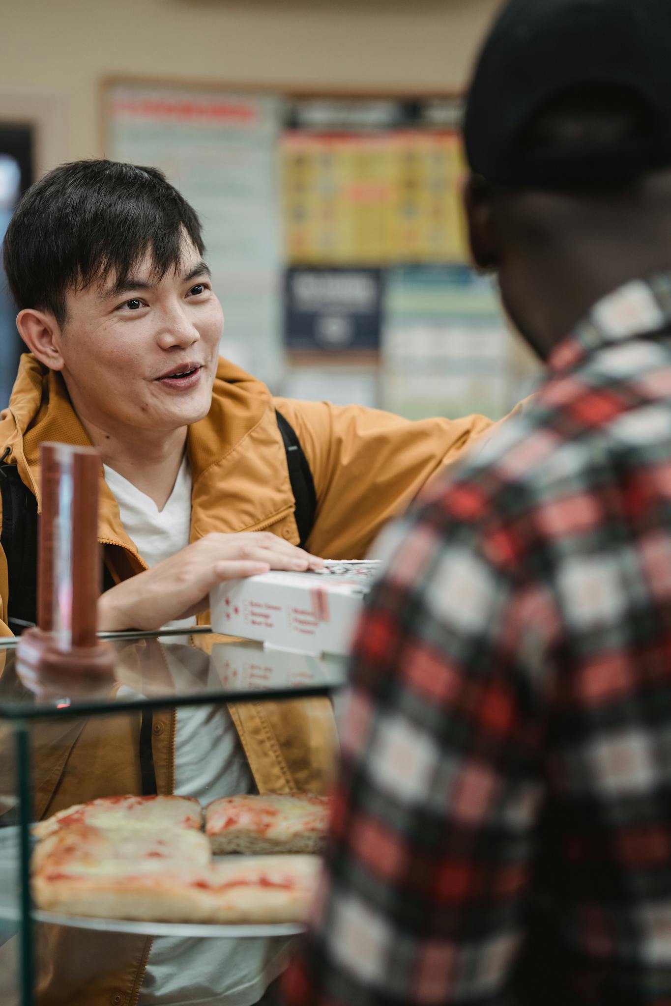 Asian man buying pizza and engaging with a cashier in a casual indoor setting.